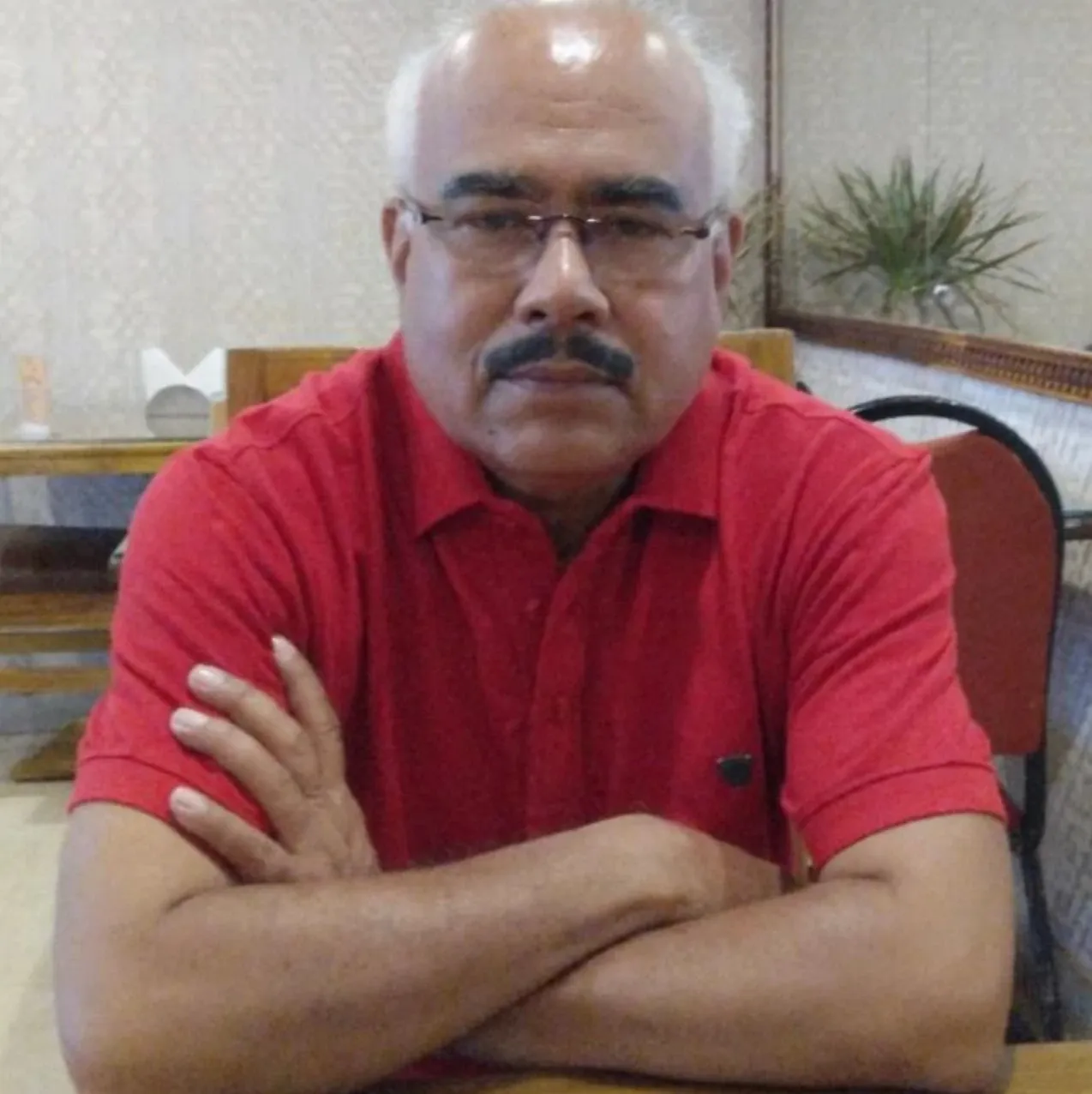Dr Bibudharanjan in glasses and a red shirt, arms folded, seated at a table.